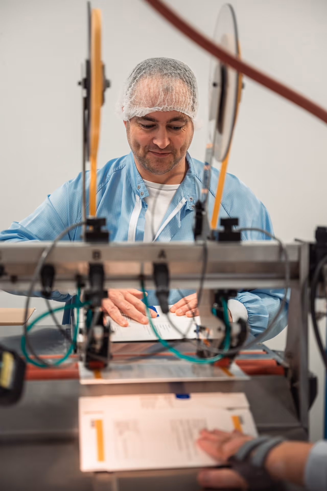 Man in a blue lab coat and hairnet works on a machine and aligns documents. He appears focused and is in an industrial or laboratory setting.