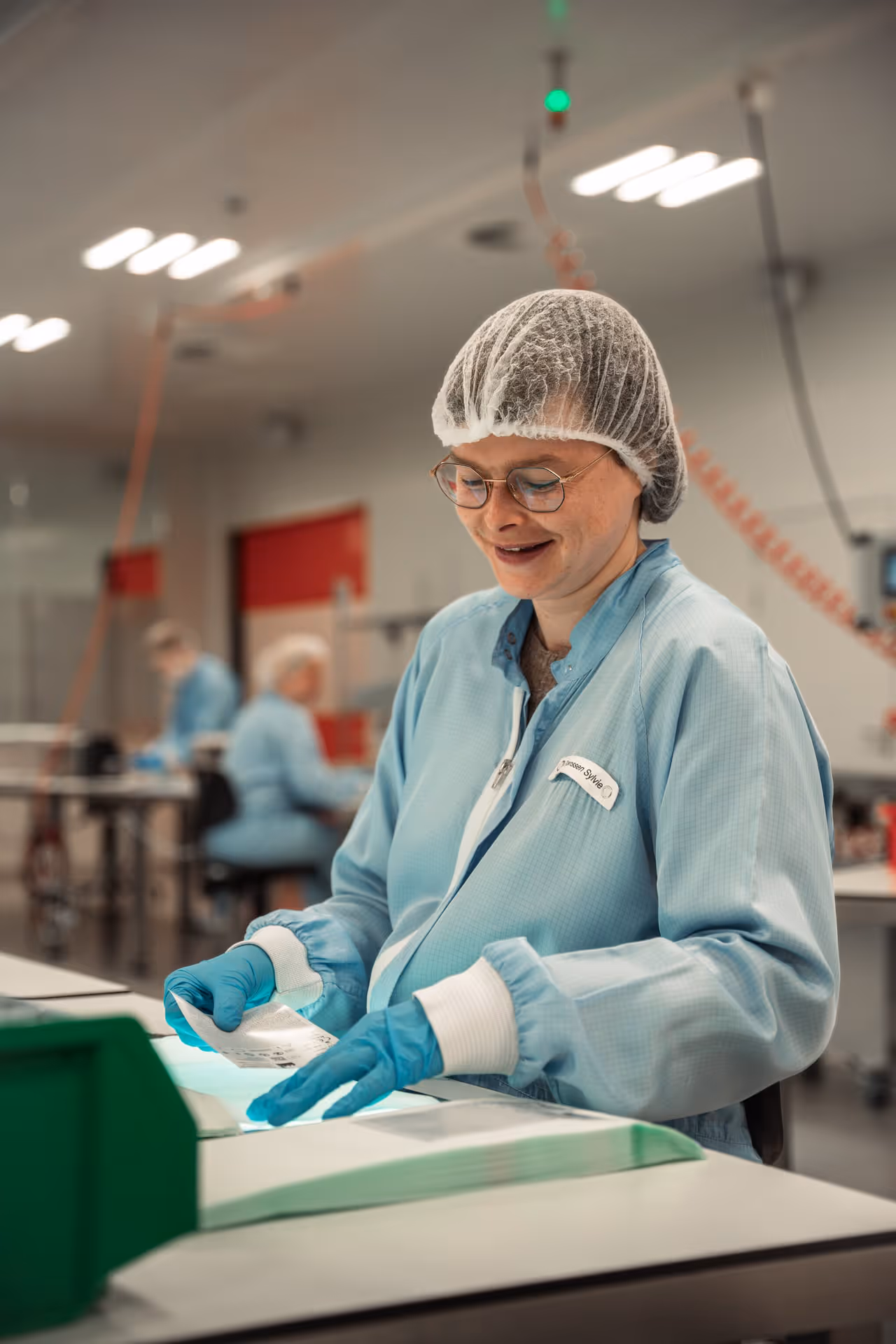 A woman wearing a hairnet and a blue lab coat smiles as she works at a table in a cleanroom. She wears gloves and handles materials with care. The room is well lit, and other employees can be seen in the background, creating a professional and focused atmosphere.