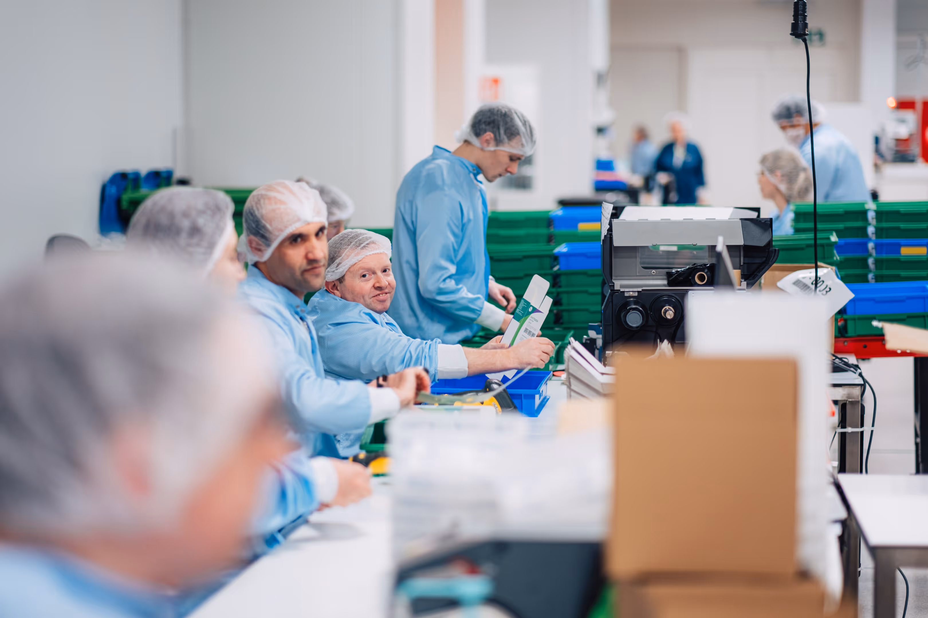 Werknemers in blauwe uniformen en haarnetjes zijn geconcentreerd bezig met assemblagetaken in een schone, georganiseerde fabriek. De sfeer is ijverig en collegiaal.