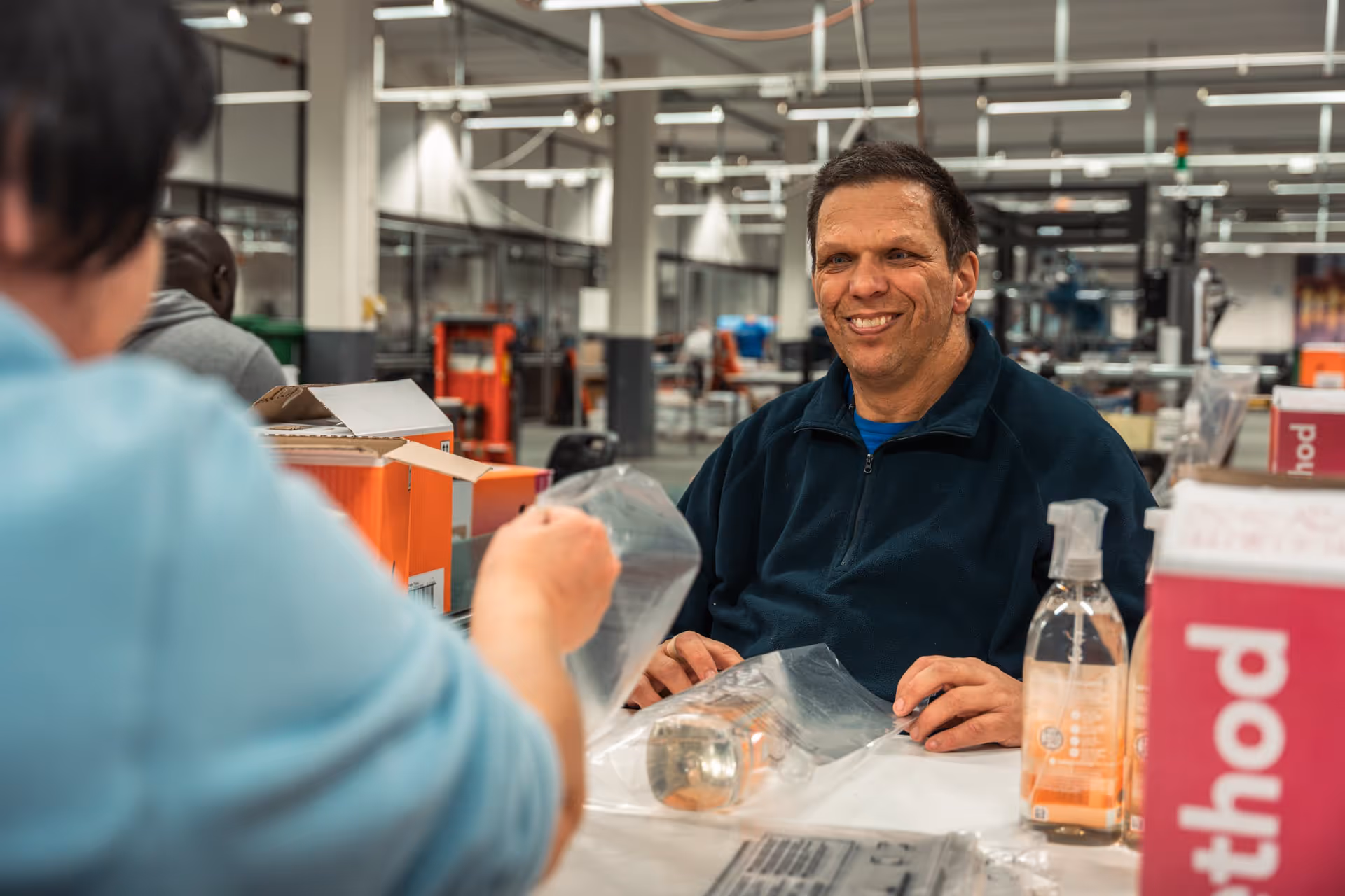 A man with a cheerful expression sits at a workstation in a busy factory and talks to a colleague. Packaging materials and bottles are on the table.
