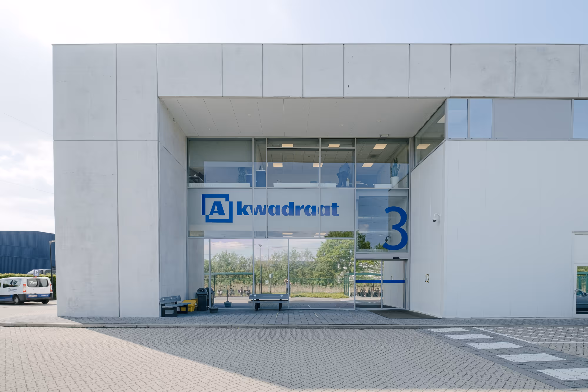 White industrial building with large glass entrance, featuring the blue logo and the text "A-kwadraat." The surroundings are characterized by a clear sky and trees.