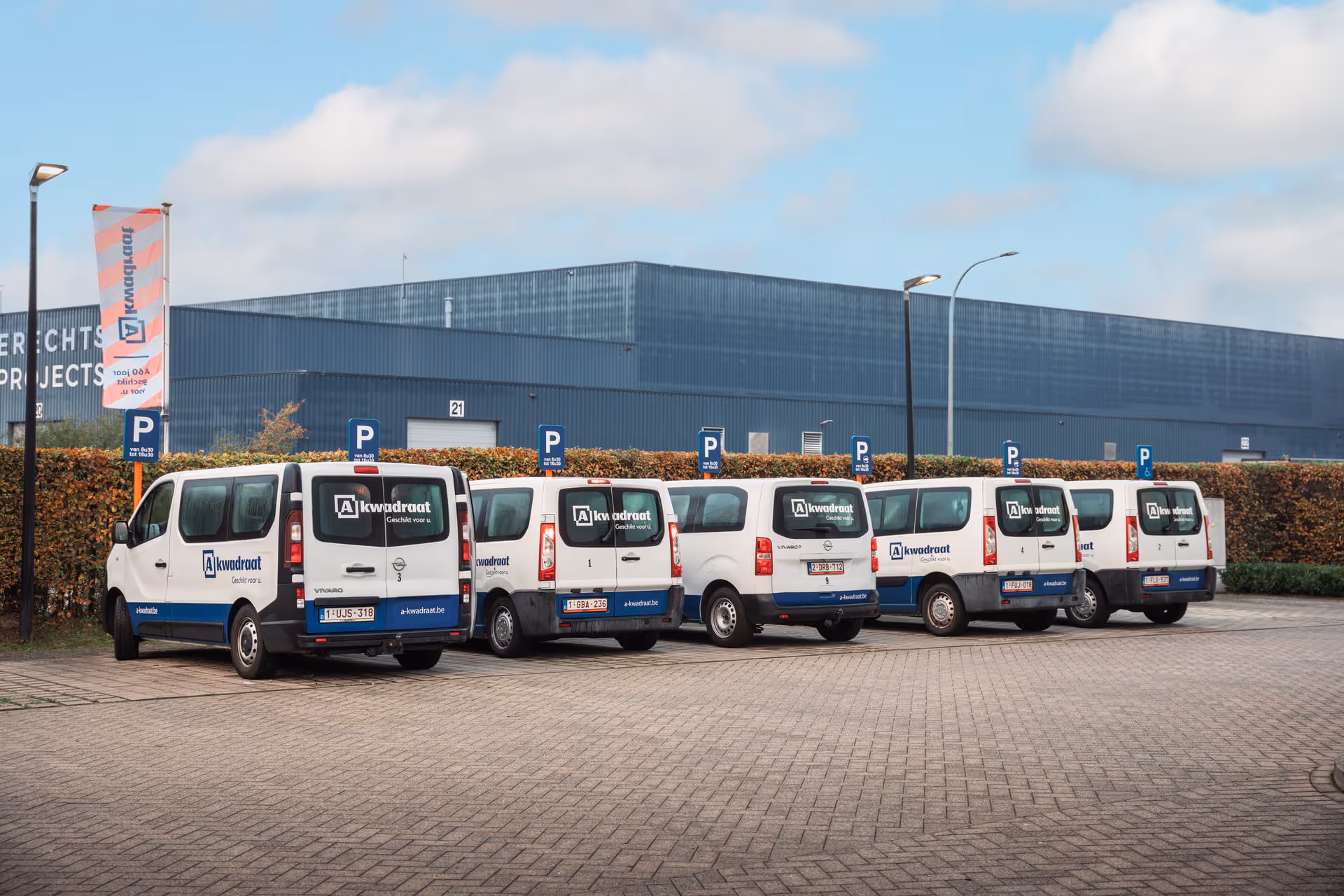 Six white and blue delivery vans with A-squared on them are parked in a row outside a large industrial building under a partly cloudy sky. The area is surrounded by hedges and parking signs.