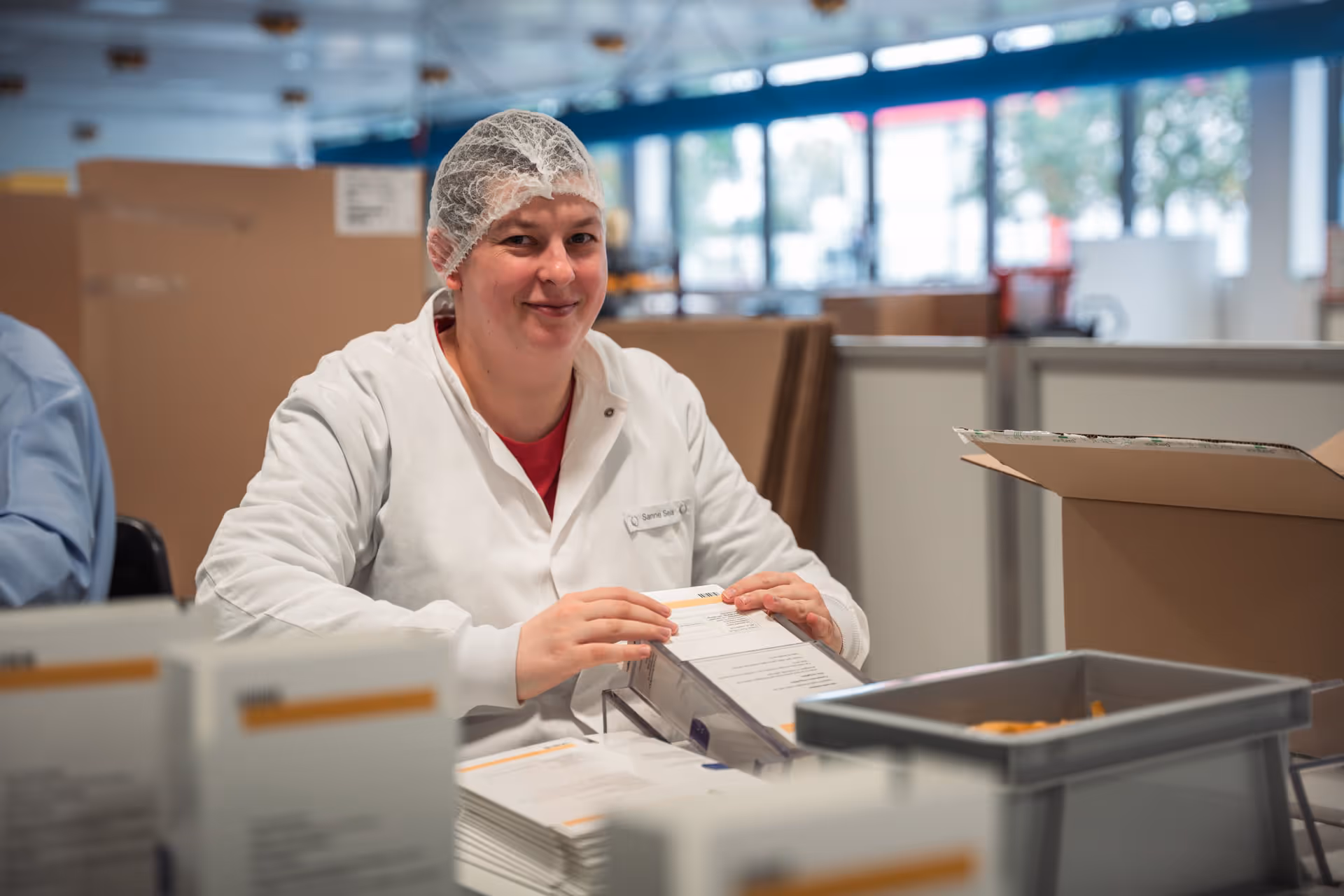 A person wearing a white lab coat and a hairnet smiles while assembling packages in a bright workspace. Open boxes and documents are scattered across the table, creating an organized and positive work environment.