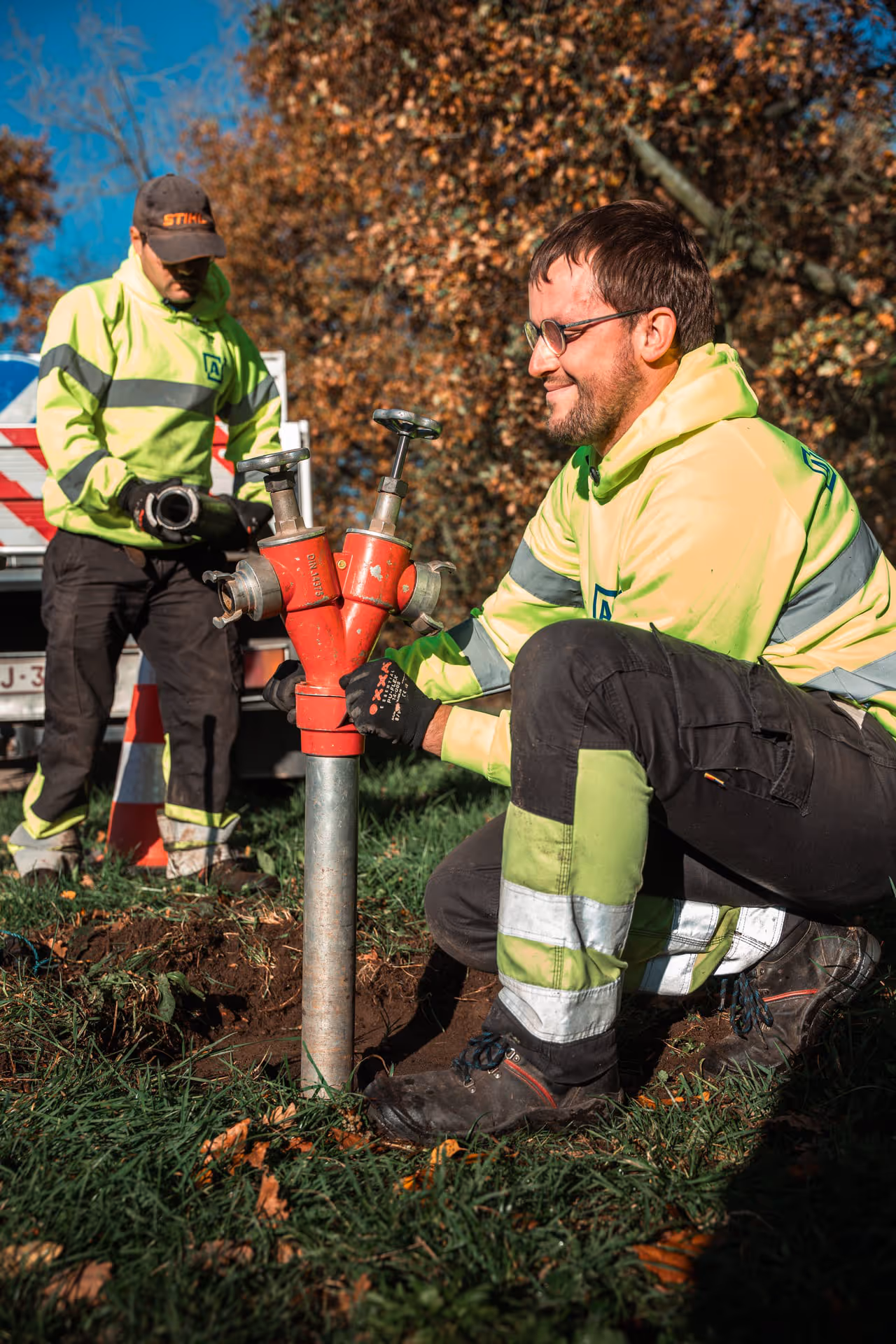 Two workers in reflective clothing are installing a red fire hydrant on a lawn. One kneels and adjusts the fire hydrant with tools, while the other assists him.
