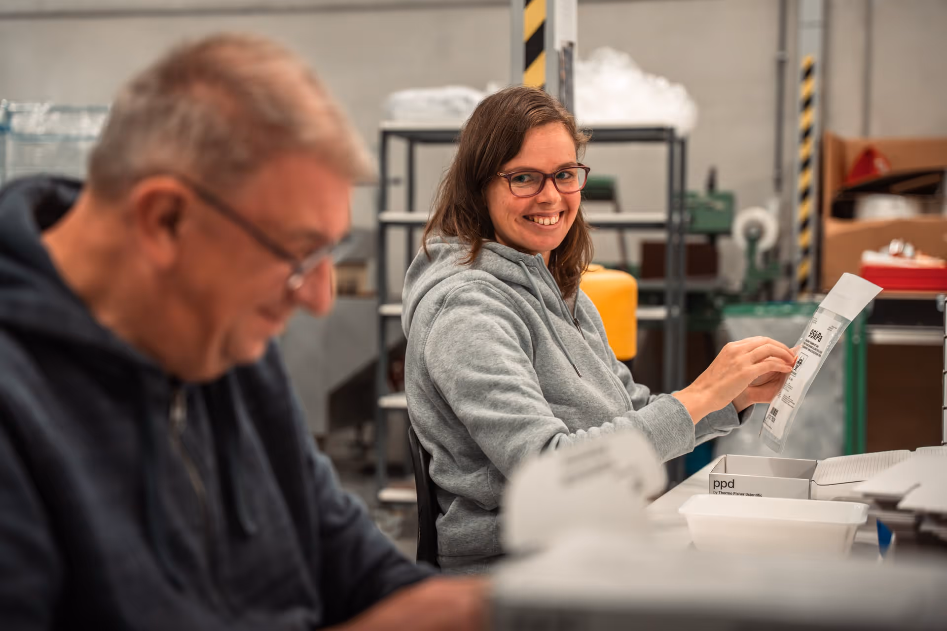 A woman wearing glasses smiles as she processes packages in a warehouse, sitting next to a blurry male colleague. The atmosphere is warm and cooperative.