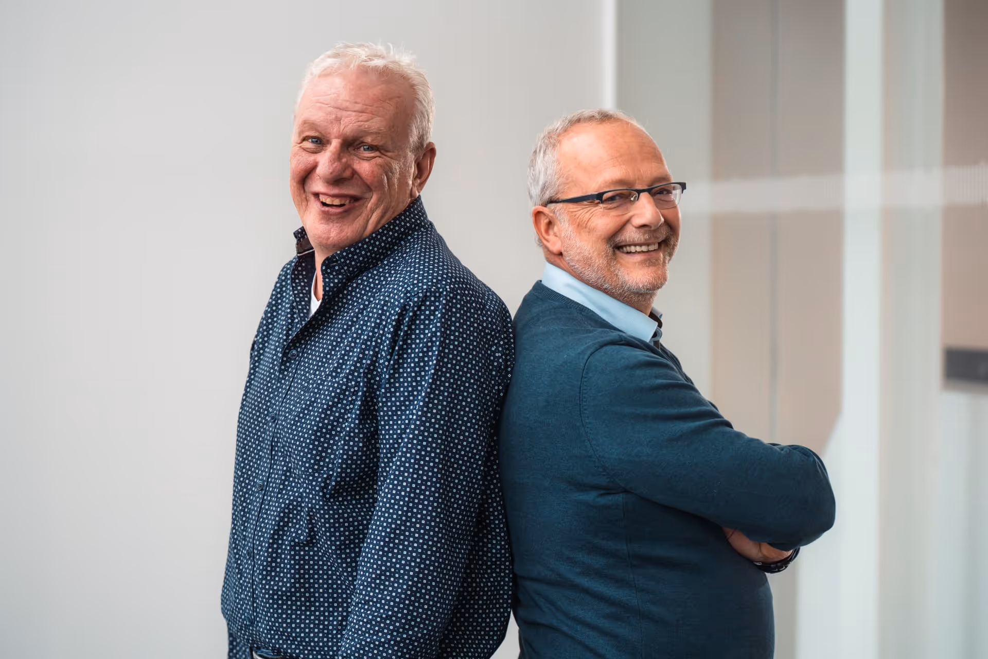Two smiling older men stand back to back in a brightly lit room. Both wear glasses and casual shirts and exude a friendly and relaxed atmosphere.