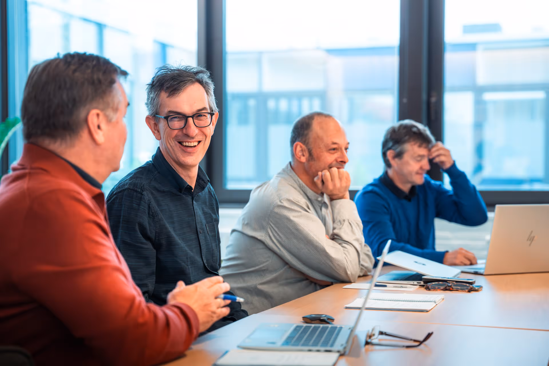 Four men are sitting at a conference table, engaged in conversation. Laptops and notebooks are open. The atmosphere is friendly, with smiles and natural light streaming in through large windows.