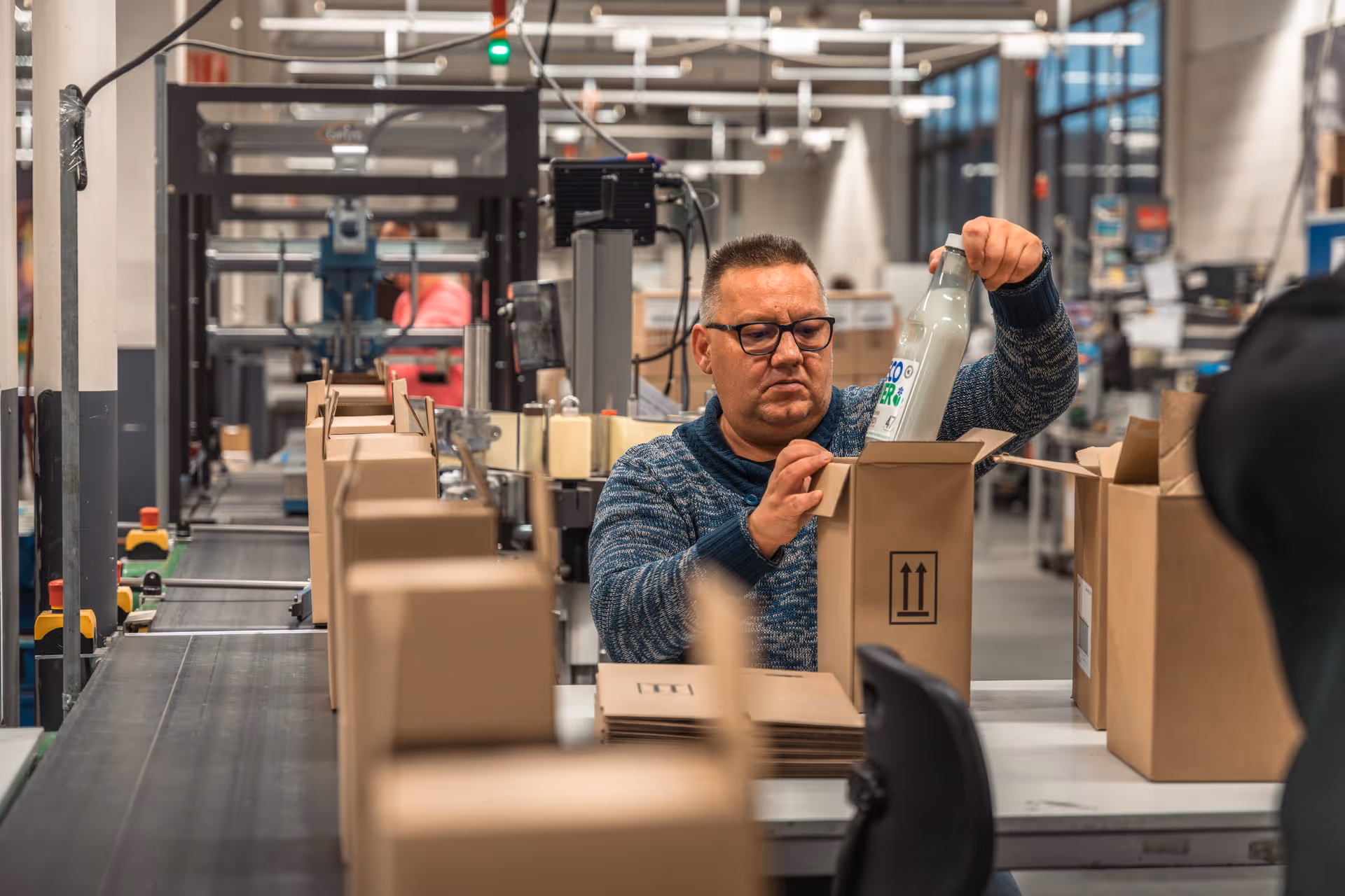 A factory worker fills cardboard boxes with bottles on a conveyor belt. The environment is industrial and exudes focus and productivity.