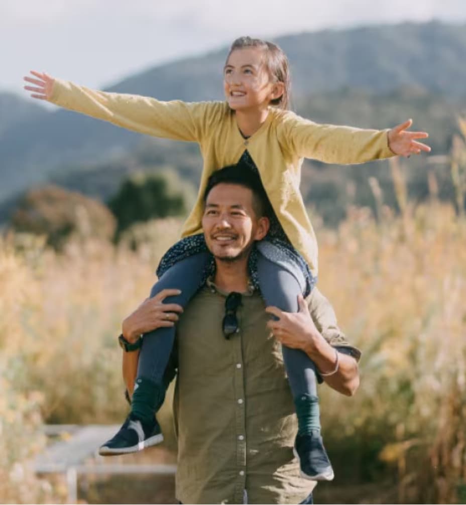 Un homme porte une fille sur ses épaules, bras étendus, dans un champ avec des montagnes en arrière-plan.