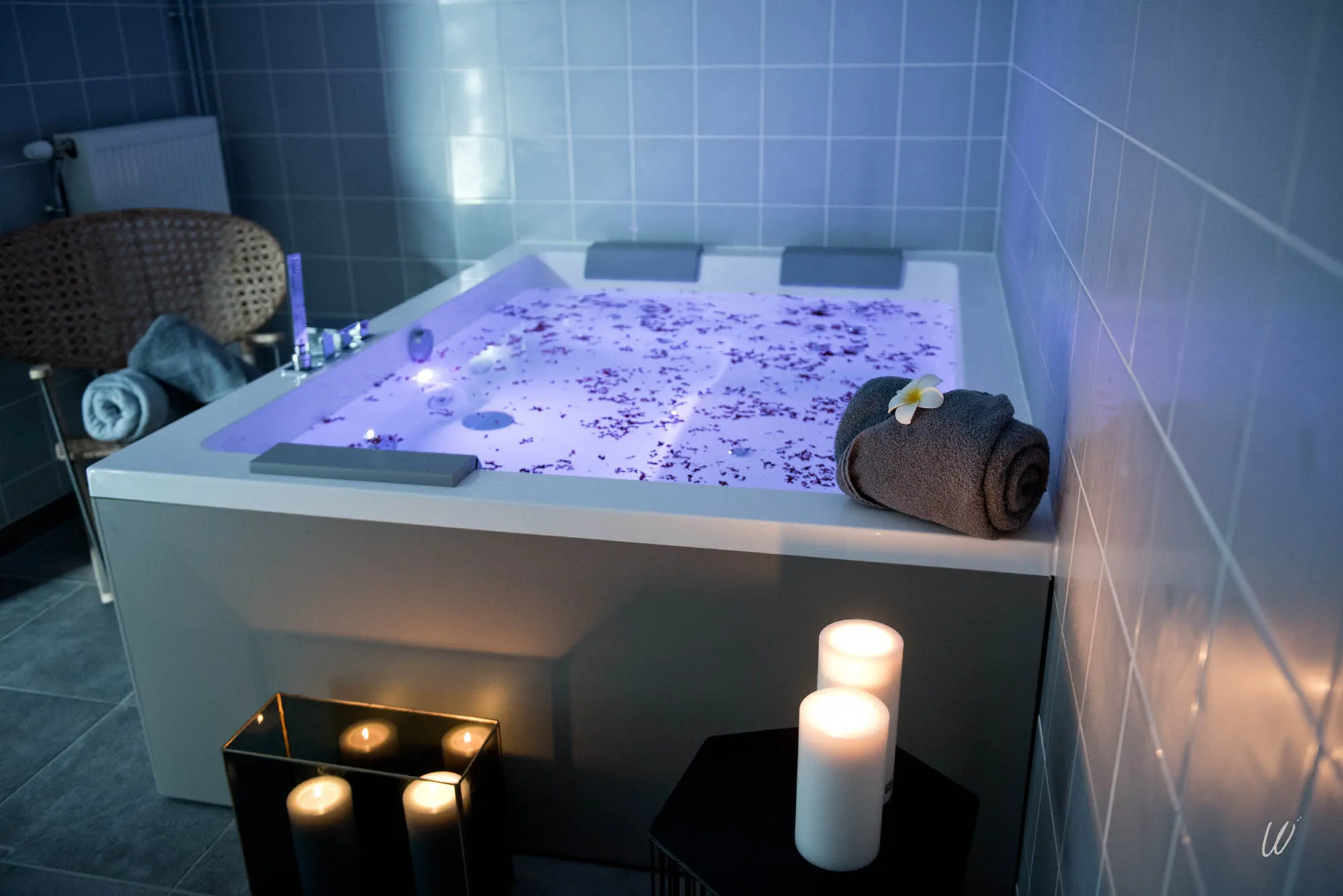 Bain jacuzzi avec eau illuminée et pétales, ambiance détente avec bougies et serviettes roulées à l’hôtel Armes de Champagne