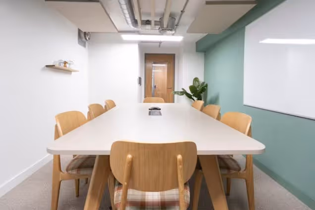 Bright meeting room with a long white table, wooden chairs with plaid upholstery, a sage green accent wall, and a large whiteboard.