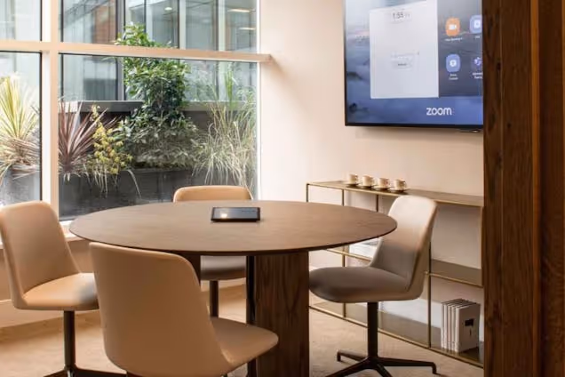 Room featuring a round wooden table, four tan swivel chairs, and a wall-mounted Zoom screen near a large window.