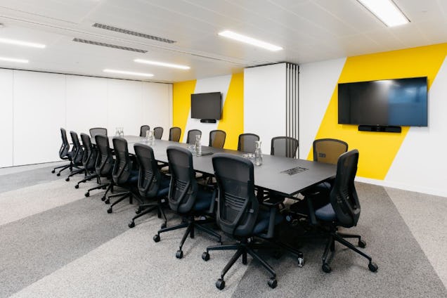 Large boardroom with a long black table and mesh swivel chairs, featuring bold yellow diagonal wall stripes and two wall-mounted screens.