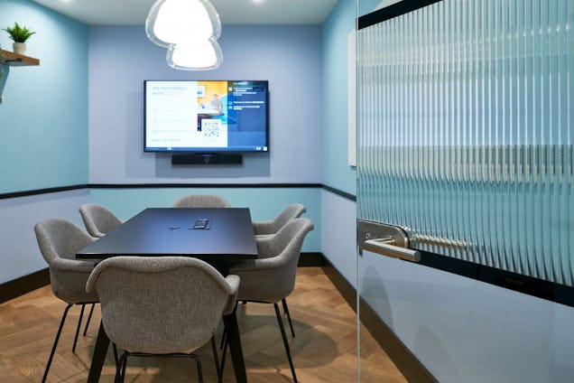 meeting room with blue-toned walls, a dark table, grey fabric chairs, and a fluted glass door in the foreground.