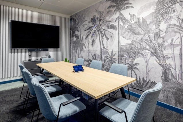 Meeting room with a light wood table, blue fabric chairs, a slatted wall with a monitor, and a grayscale tropical mural.