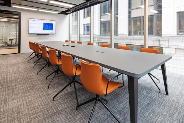 Boardroom with a long grey table and orange swivel chairs, featuring floor-to-ceiling windows overlooking city buildings.