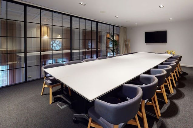 Large conference room with a long white table, grey tub chairs, and a black-framed internal glass wall.