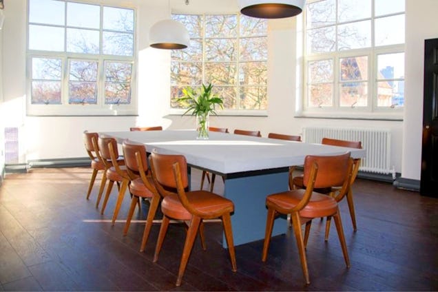 Room with large windows, a long white table and several wooden chairs surrounding it