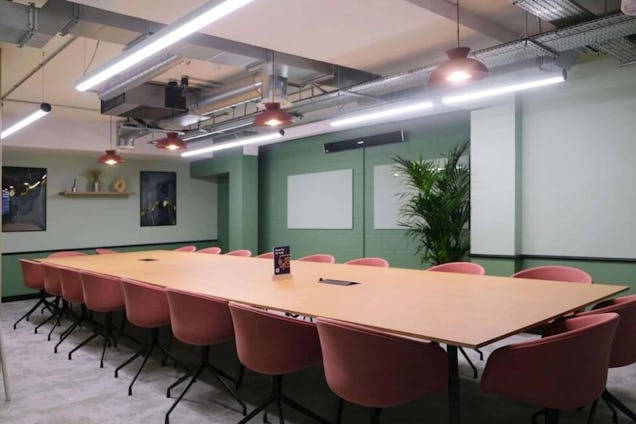 Space with a long wooden table and pink lush chairs around it. The walls are different shades of green and their are pink hanging lights.