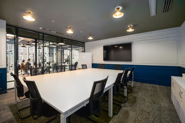 White table with black long chairs surrounding it. The walls are dark blue and white and there is a TV on the wall, with a whiteboard beside it