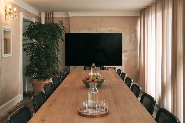 Warmly lit room with beige/pink walls and curtains. There's a long wooden table with black chairs surrounding it. A fruit bowl is on the table and glasses of water.