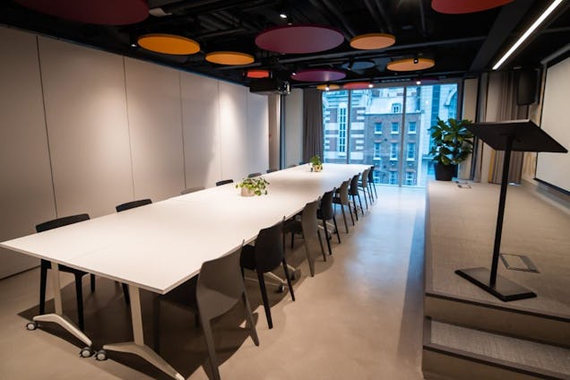 Room with a large white table surrounded by black and grey chairs. There is a small stage with a display screen and a stand for papers. There are different colour circle decor on the ceiling