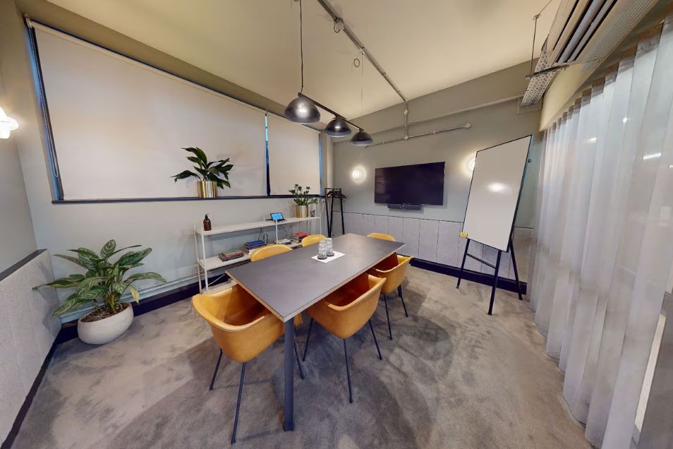 Space with a small dark wood table with yellow box chairs. There are silver hanging lights, a green plant in a white pot, a TV and a large whiteboard