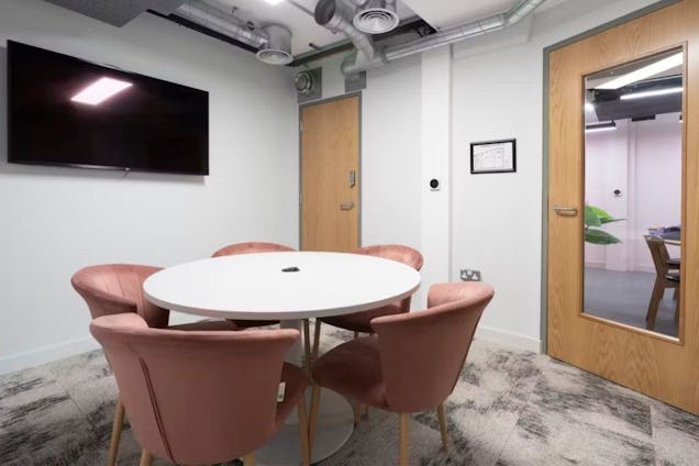 Small meeting room featuring a white round table, dusty pink velvet chairs, a wall-mounted TV, and industrial exposed ceiling vents.
