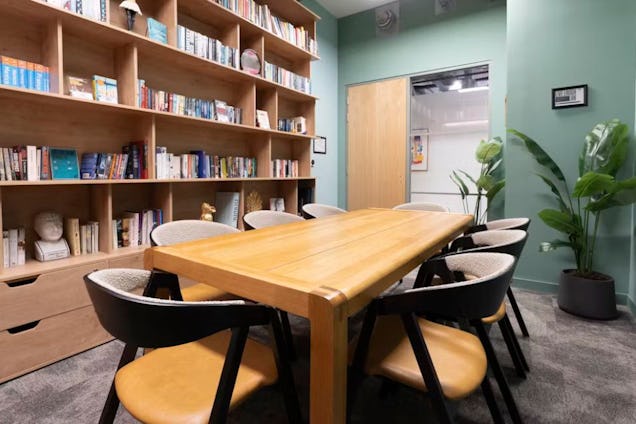 Library-style meeting room with a large wooden table, yellow-seated chairs, and a floor-to-ceiling bookshelf against a sage green wall.