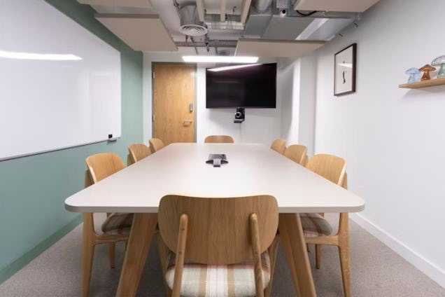 Modern meeting room with a long white table, wooden chairs with plaid seats, a sage green accent wall, and a large whiteboard.