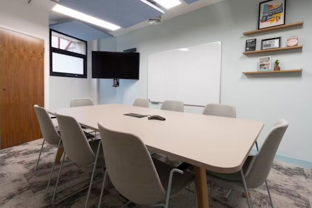 Meeting room with a light grey table, matching fabric chairs, floating wooden shelves, and a large whiteboard on a pale blue wall.