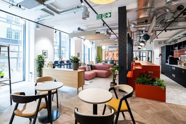 Bright, open-plan industrial office featuring herringbone wood floors, a pink sectional sofa, cafe-style seating, and exposed ceiling ductwork.