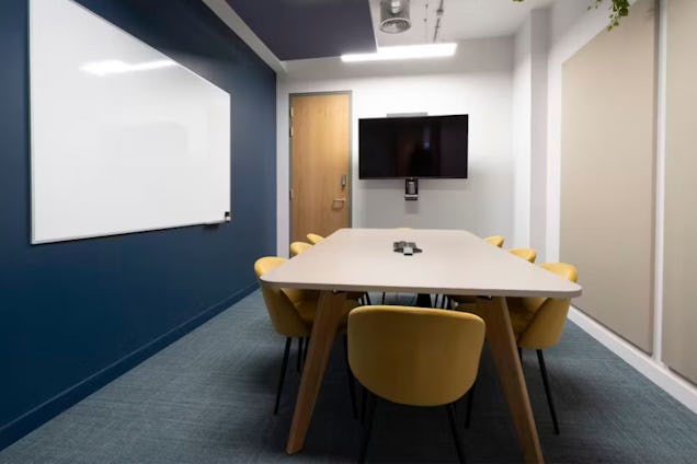 Meeting room with a long white table, yellow chairs, a navy blue accent wall with a whiteboard, and a wall-mounted TV.