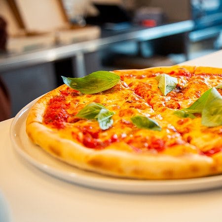 Close-up of a fresh Margherita pizza with melted cheese and basil leaves on a white plate, set against a blurred kitchen background.