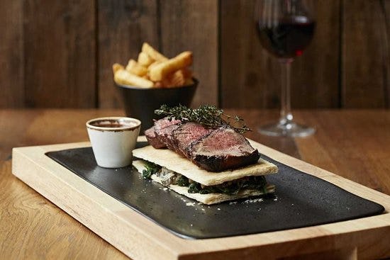 Slices of medium-rare steak garnished with herbs, served atop a layered crisp with greens, accompanied by fries and red wine.