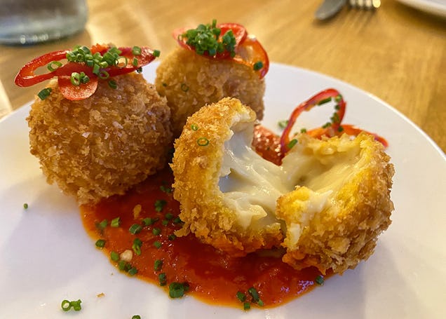 Fried arancini balls served over tomato sauce, garnished with red chilli rings and chives, with one ball broken open to reveal melted cheese.