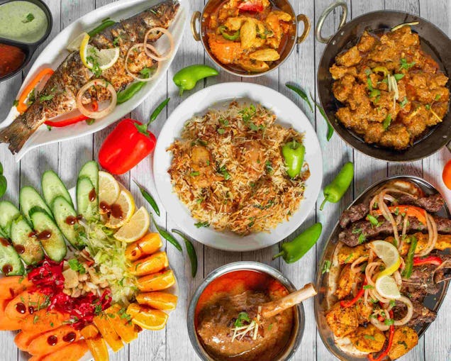 Overhead view of a South Asian food spread with biryani, grilled fish, kebabs, lamb shank curry, and a vibrant vegetable salad.