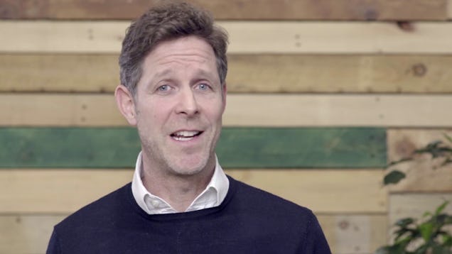 A man in a dark sweater over a white collared shirt speaks in front of a horizontal wood-panelled wall.