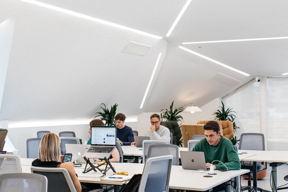 Minimalist office with a sloped white ceiling and recessed linear lighting, where people work at long white communal desks.