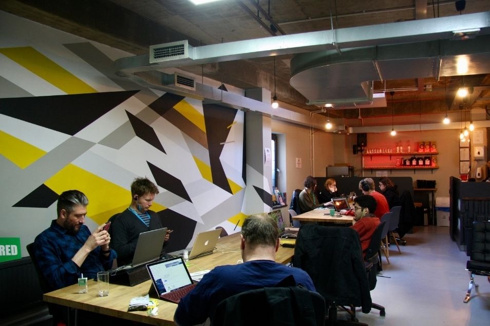 Coworking space with a bold geometric mural in yellow, black, and white, featuring people working at long wooden tables.