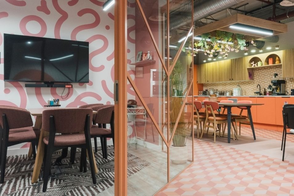Vibrant office break room featuring pink patterned wallpaper, orange kitchen cabinetry, and a glass partition separating a dining area.