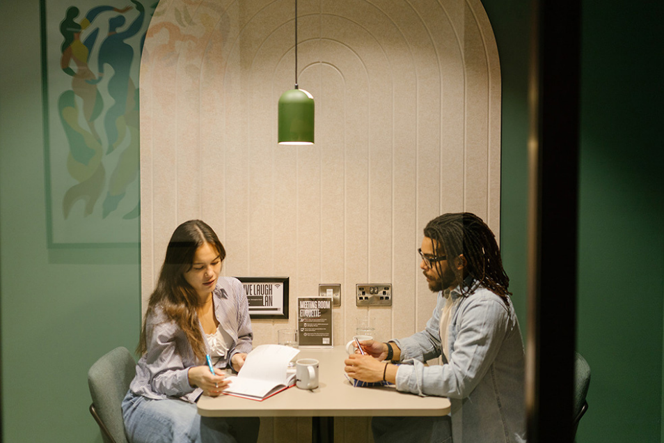 Two people sit at a small table in a modern booth with an arched back, focused on paperwork under a green pendant light.