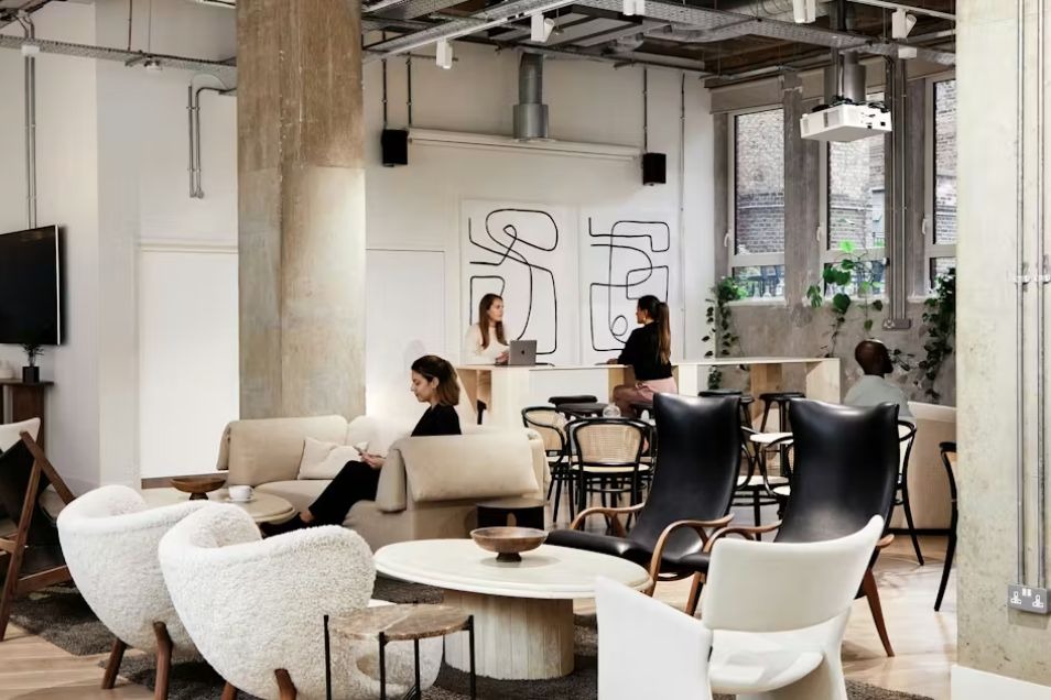 A chic, open-plan office lounge featuring cream bouclé and black leather armchairs, industrial concrete pillars, and people working in a bright, modern setting.
