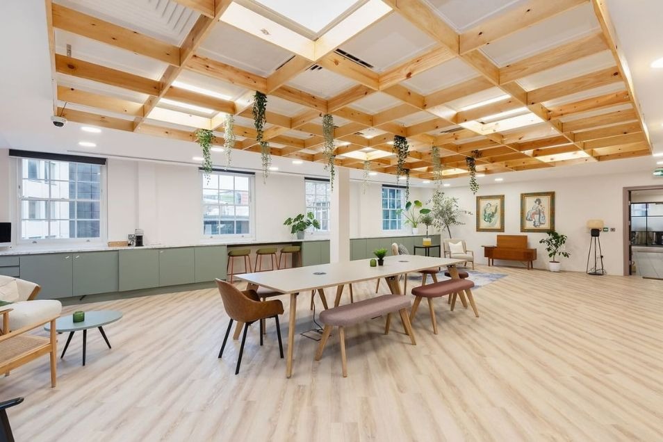 A spacious, light-filled office breakout area featuring a large wooden lattice ceiling with trailing plants, sage green cabinetry, and communal dining tables.