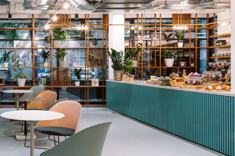 Office café featuring a teal ribbed counter, pastel chairs, and a floor-to-ceiling wooden lattice shelf filled with indoor plants.