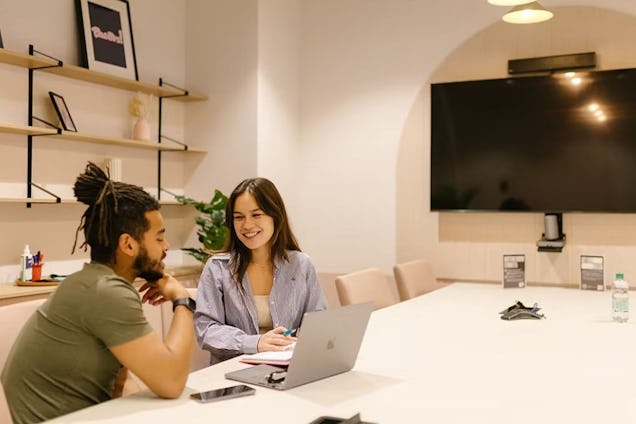 Two people sit at a white table and talk in front of a laptop in a room with shelves and a television.