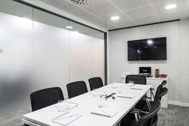 A white table with black chairs and notebooks in a meeting room featuring frosted glass walls and a television.