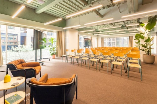 Rows of chairs face two armchairs and a table in an event space with windows, plants, and overhead lights.