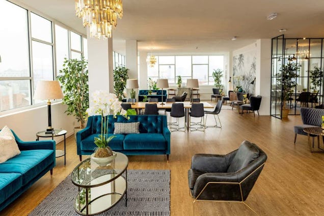 A lounge area with blue sofas, leather chairs, and a wooden table in an office space with large windows and chandeliers.