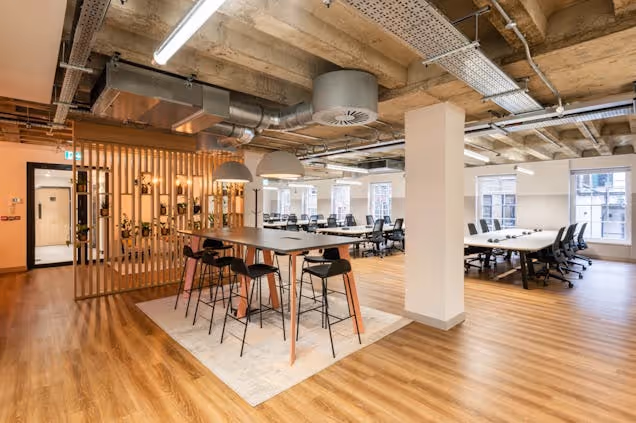 A photo of the co working space, large tables and stools, then desks and office chairs in the background.
